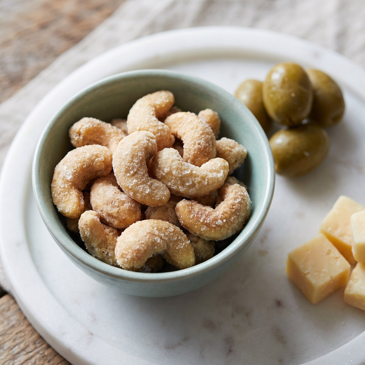 Cajous au sucre d'érable croustillants dans un bol vert, servis sur un plateau de marbre avec des olives et du fromage