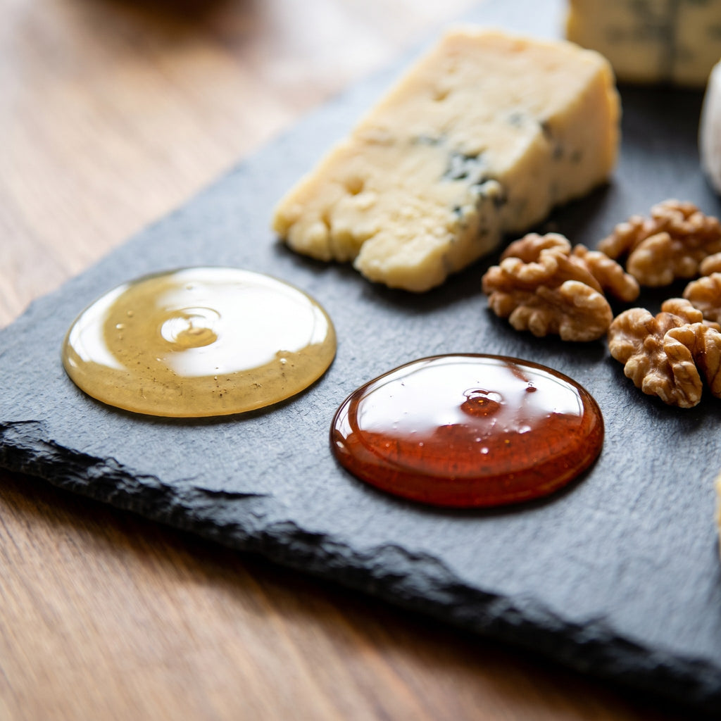 Duo de miel doré et ambré présenté sur une ardoise noire à côté de morceaux de fromage bleu et de quelques noix