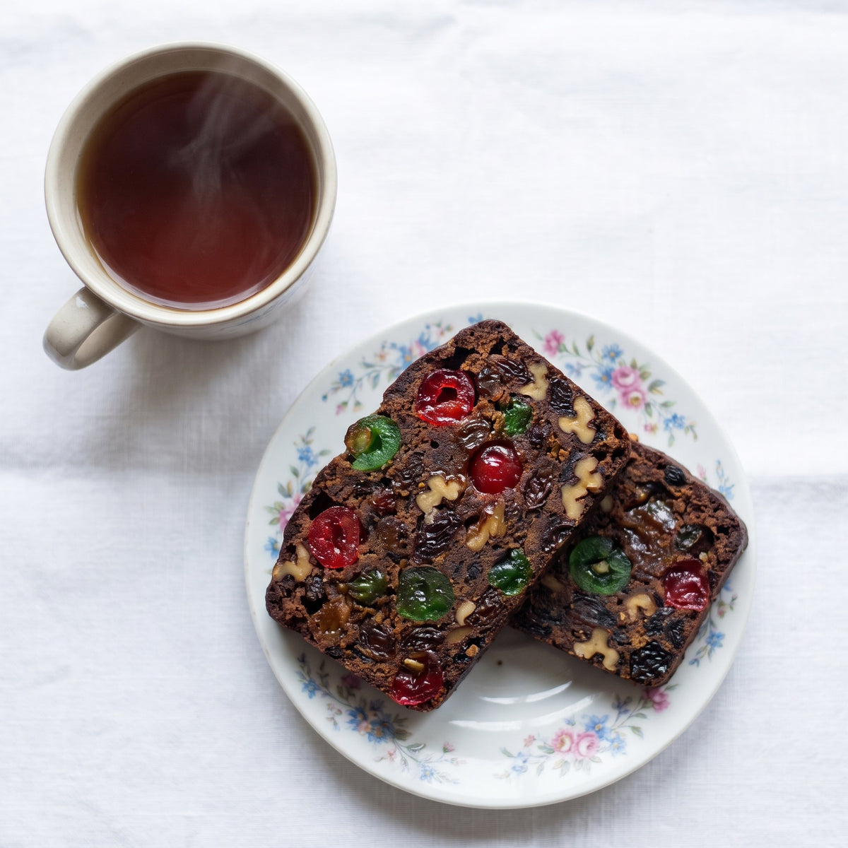 Deux tranches de gâteau aux fruits garnies de cerises confites, servies sur une assiette fleurie à côté d'un thé chaud