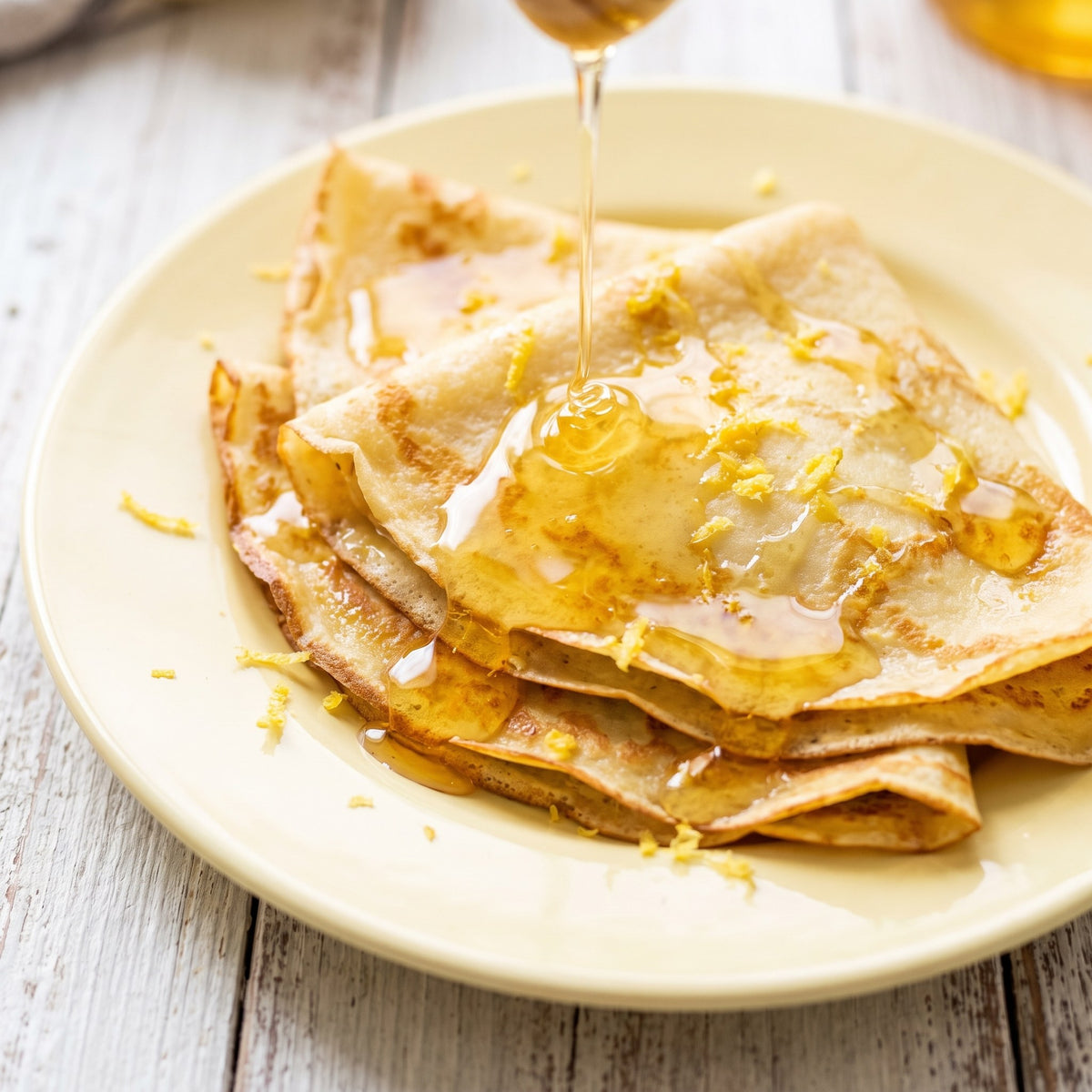 Crêpes pliées sur une assiette jaune, arrosées d'un filet de Miel du printemps et parsemées de zestes de citron