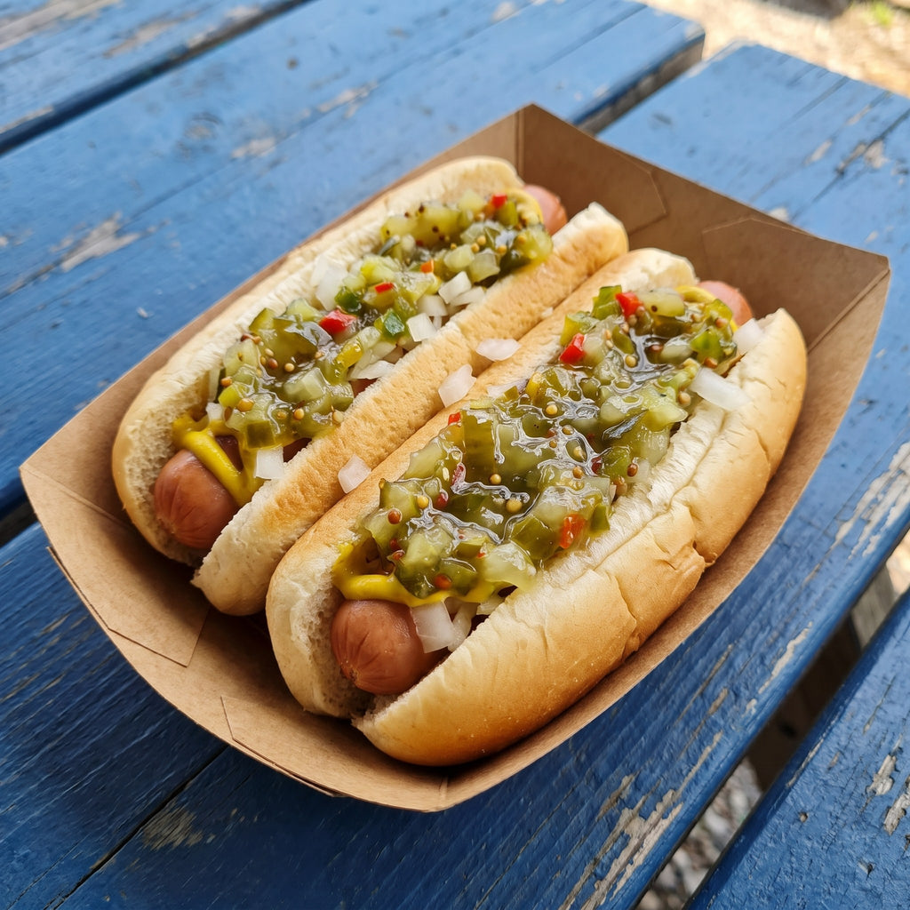Deux hot-dogs garnis de relish, moutarde et oignons blancs, servis dans un petit plateau sur une table bleue usée