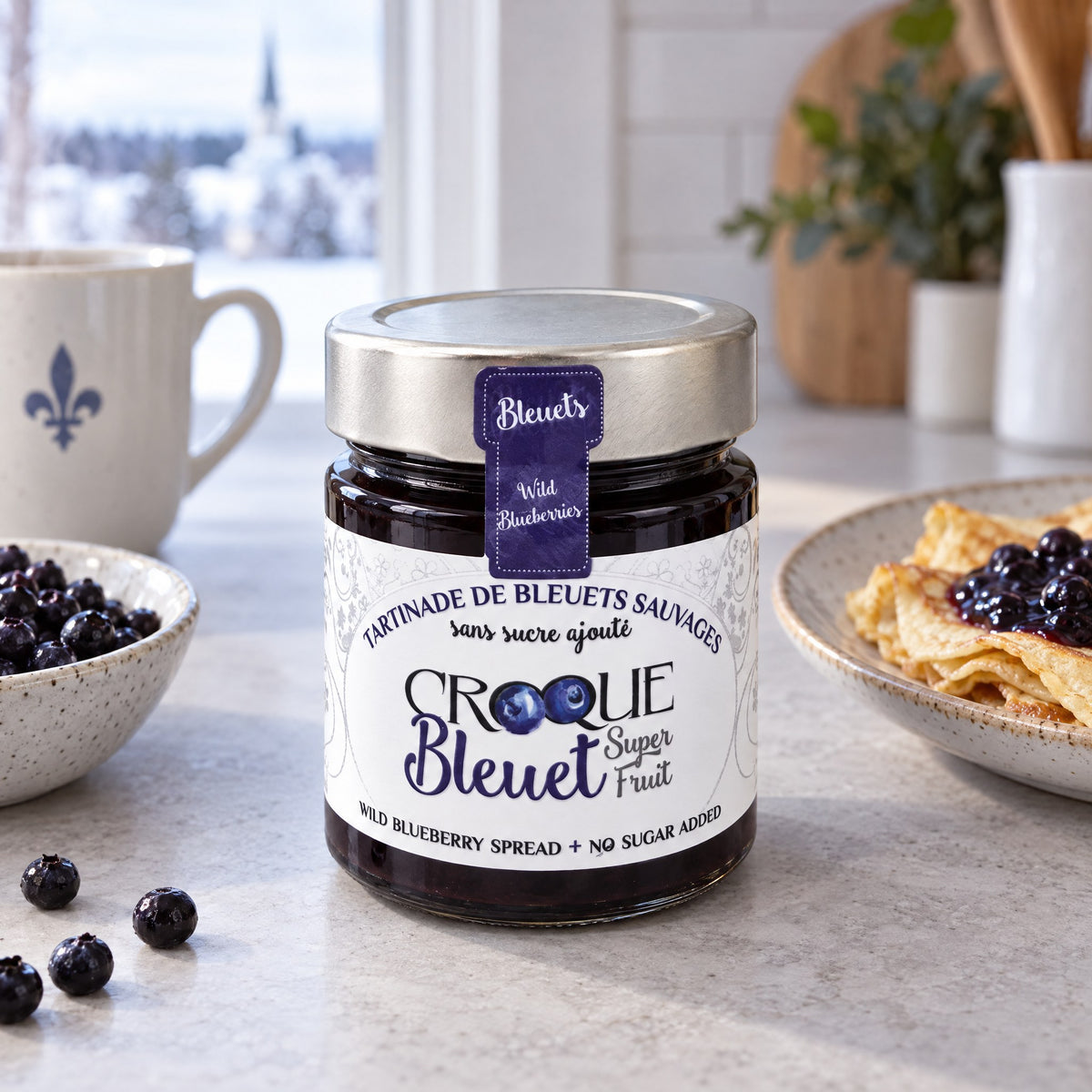 Pot de tartinade Croque bleuet au centre d’une table, avec bleuets frais, crêpes garnies et tasse blanche