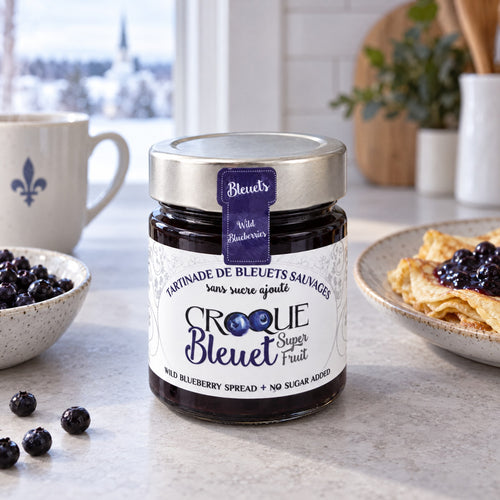 Pot de tartinade Croque bleuet au centre d’une table, avec bleuets frais, crêpes garnies et tasse blanche