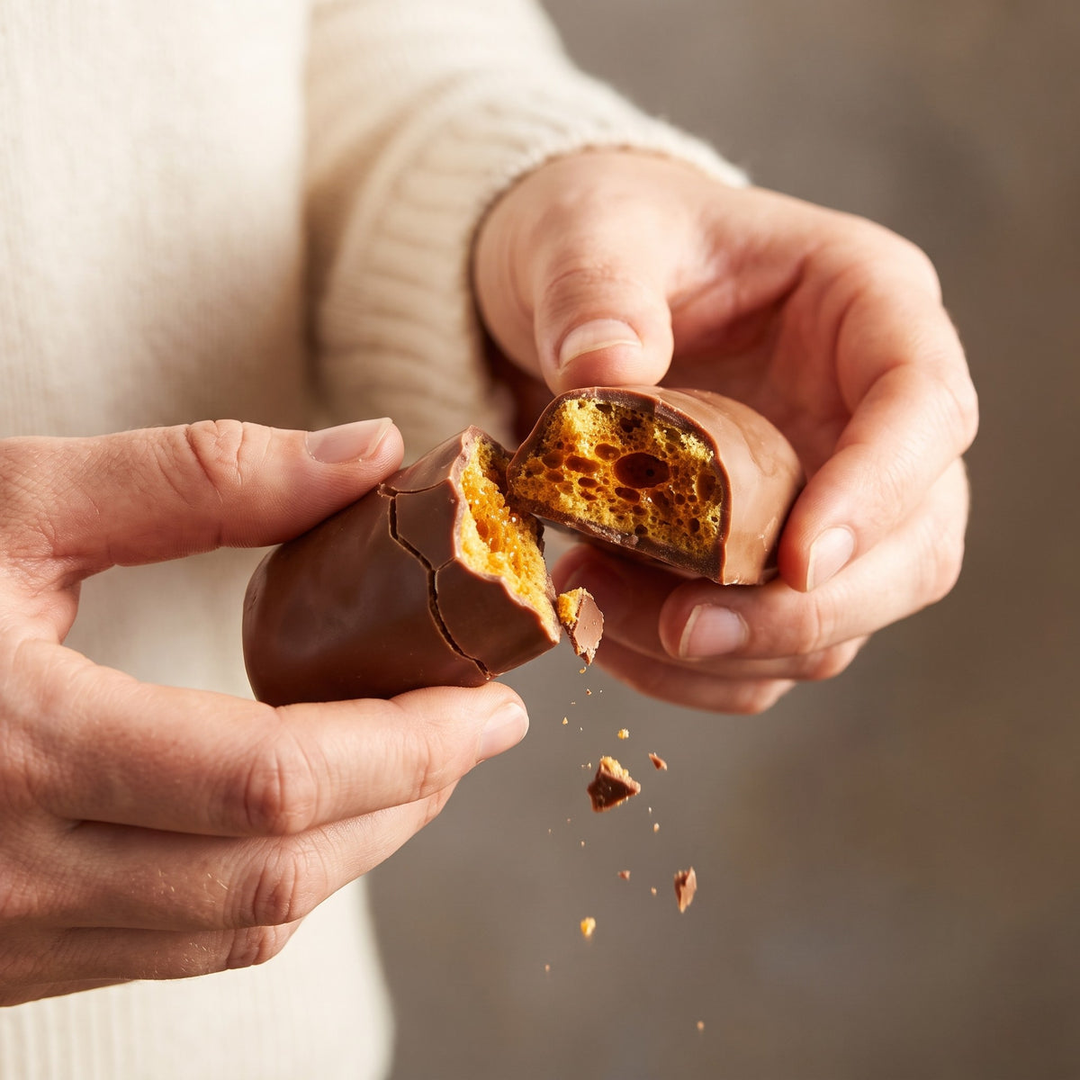 Deux mains cassent une tire éponge enrobée de chocolat, révélant un cœur alvéolé doré